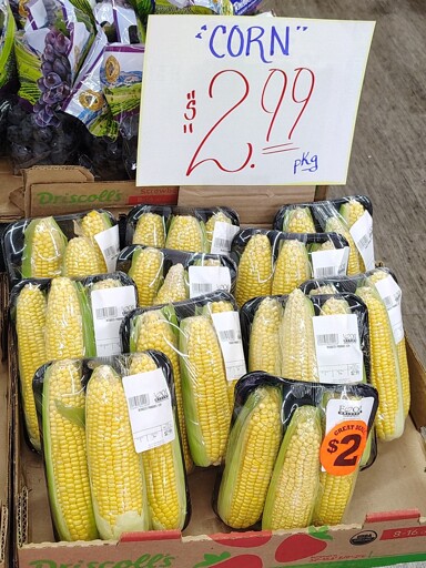 Display of packaged ears of corn at a grocery store. The display is labeled with a handwritten sign reading "corn $2.99 per package" but the word "corn" is in unnecessary quotation marks.