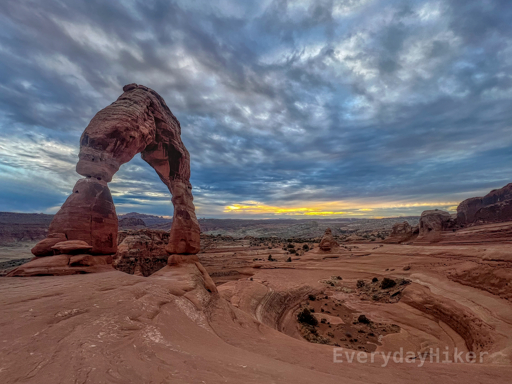 Delicate Arch and its forum-like bowl beside it, as the setting sun breeches the thick clouds along the horizon.
